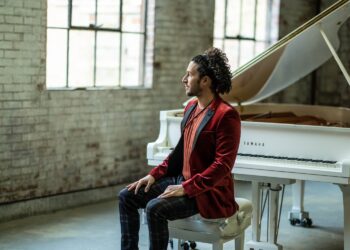Pianist, Emmet Cohen sitting on a piano bench looking to the left.